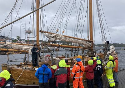 Folk i fullt seilutstyr med redningsdrakt og regntøy, står på linje foran en treseglbåt.
