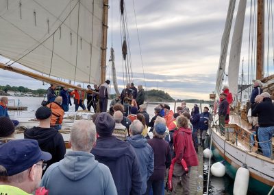 En folkemengde står foran en treseilbåt med seglene oppe.