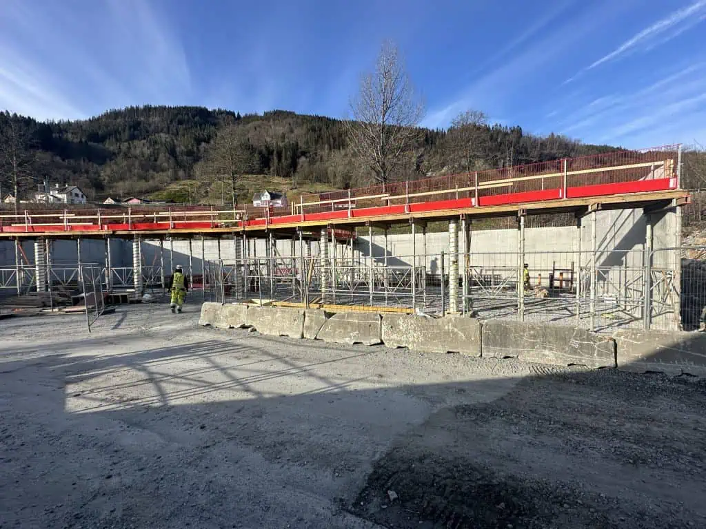 Blå himmel over en anleggsplass, store betongvegger er satt opp bak et byggegjerde. En heiskran. Bakker og skog i bakgrunnen