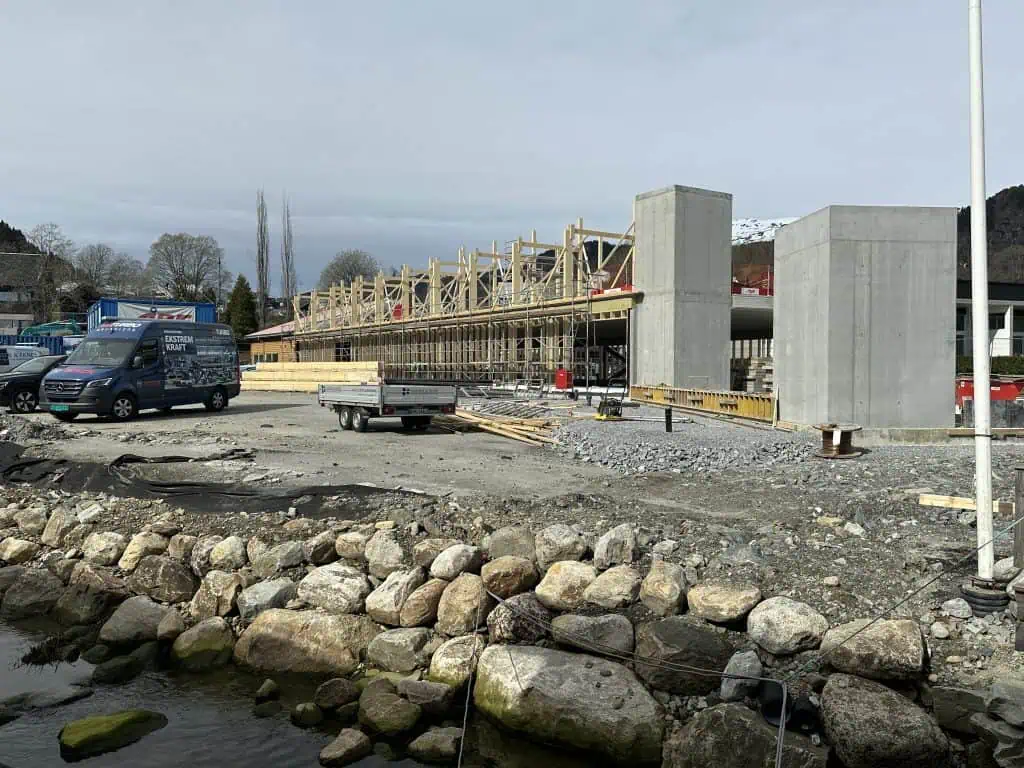 alt="Et nytt bygg skrider frem. En gul trebygning står inntil et nybygg som bare består av betongskiller og stålbjelker og en heissjakt. I bakgrunnen er det fjell og fjord.