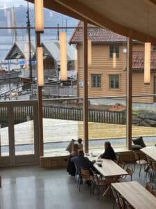 Kafe med utsikt over båter og fjord. Det sitter tre personer ved et langbord og ser ut det store vinduet som dekker det meste av veggen.