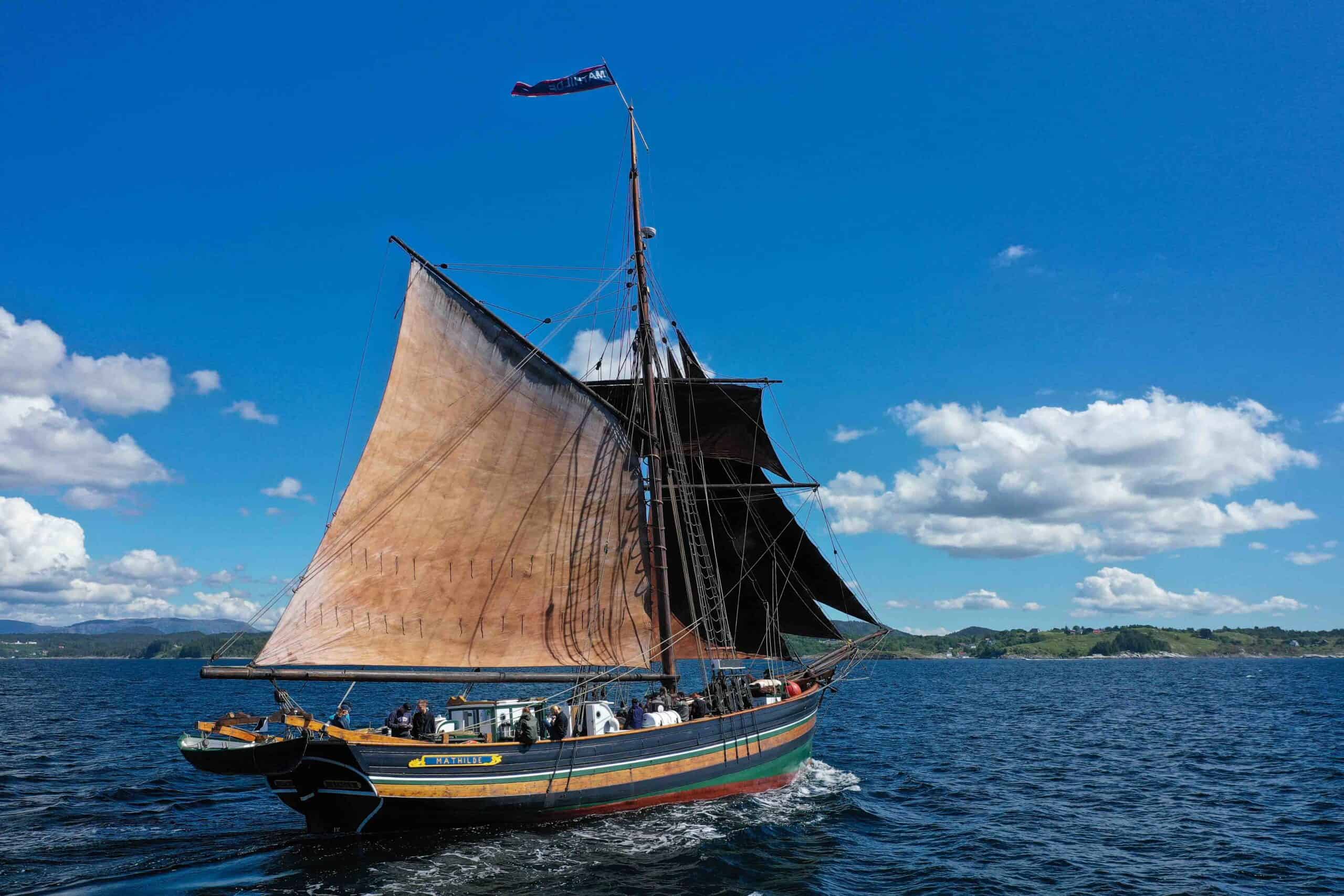 Mathidle seglar på på fjorden, med grøne bakkar og blå himmel i bakgrunnen.